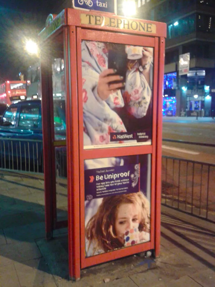 Red telephone booth with mismatched advertisement panels at night.