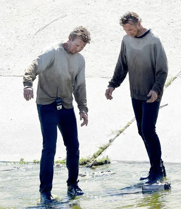 Two men dressed alike in dirty sweatshirts and dark pants standing in shallow water, showcasing celebrity body doubles.
