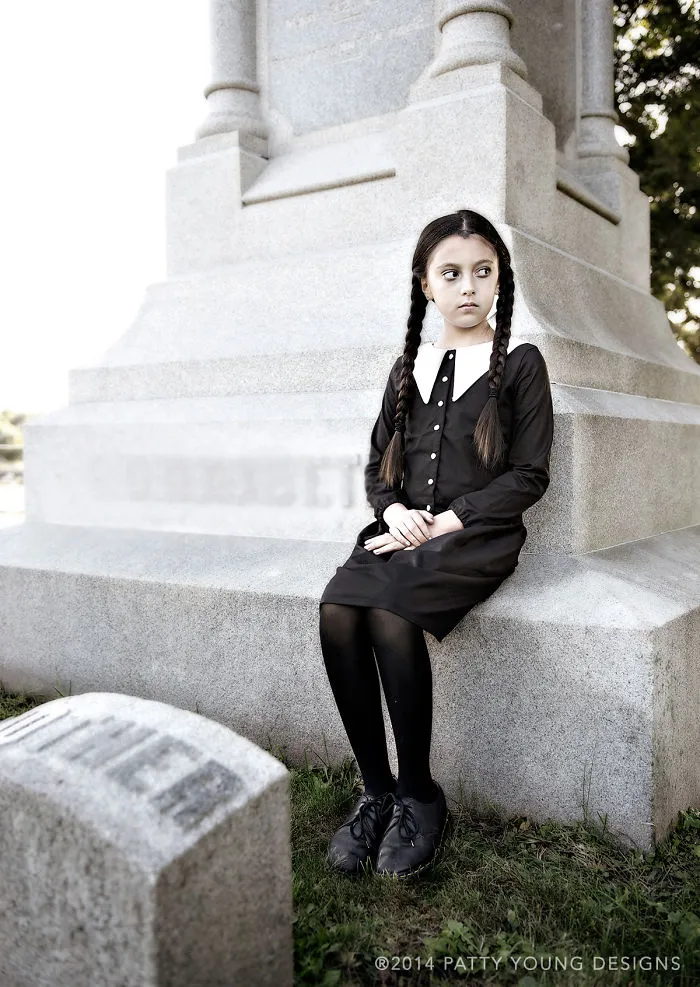 A girl dressed as Wednesday Addams sitting near a tombstone for creative Halloween costume ideas