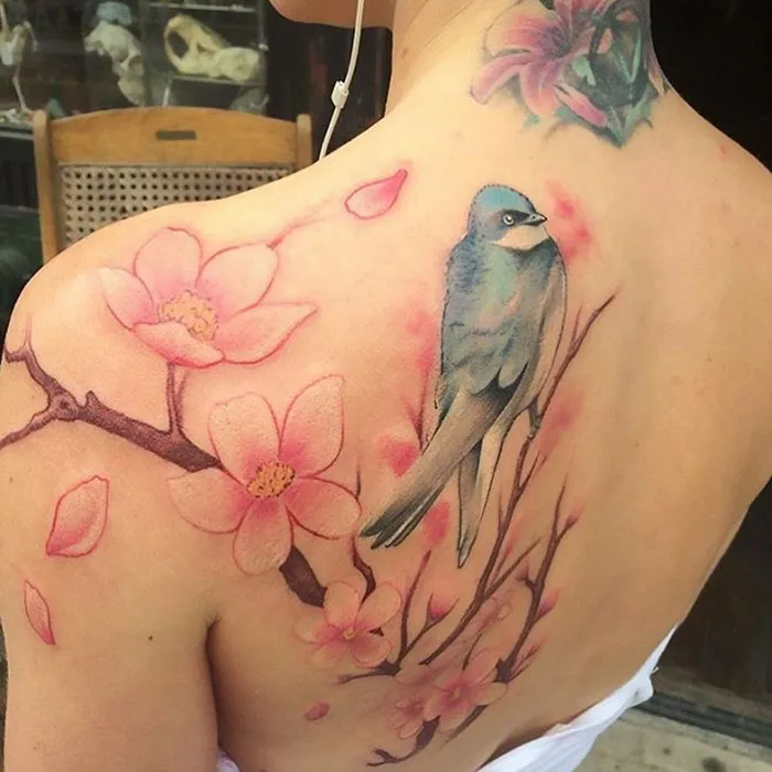 Small bird tattoo on back with pink cherry blossoms and watercolor design.