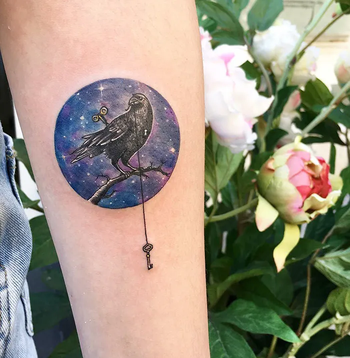 Small bird tattoo featuring a cosmic background and a key, inked on forearm near flowers.