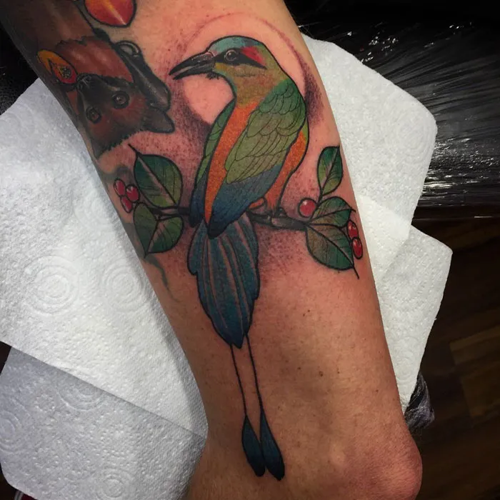 Colorful small bird tattoo with green wings and orange accents on a branch, inked on an arm.