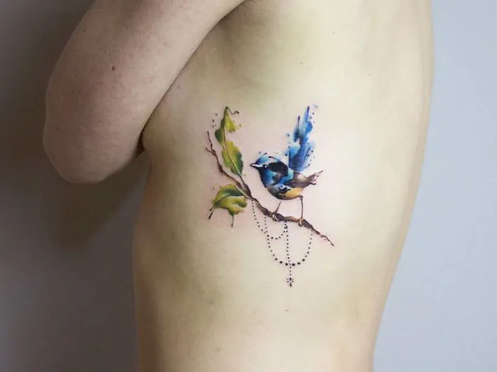 Colorful small bird tattoo on skin with green leaves and a delicate branch.