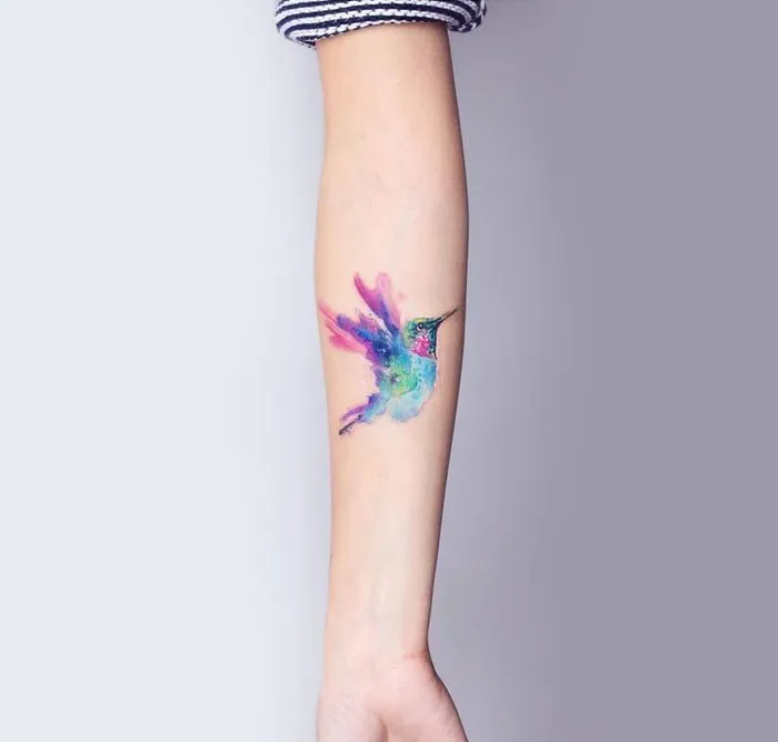 Colorful small bird tattoo on forearm, vibrant watercolor design in purple, blue, and green tones.