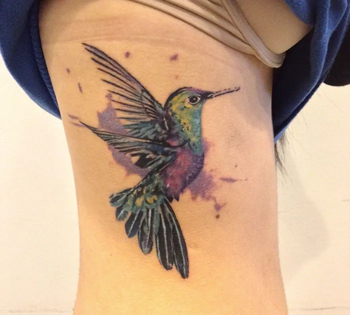 Colorful small bird tattoo on skin, with watercolor effect, featuring vibrant shades of blue, green, and purple.