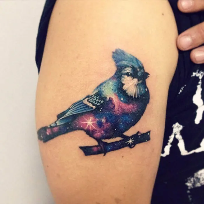 Colorful small bird tattoo with galaxy design on upper arm.