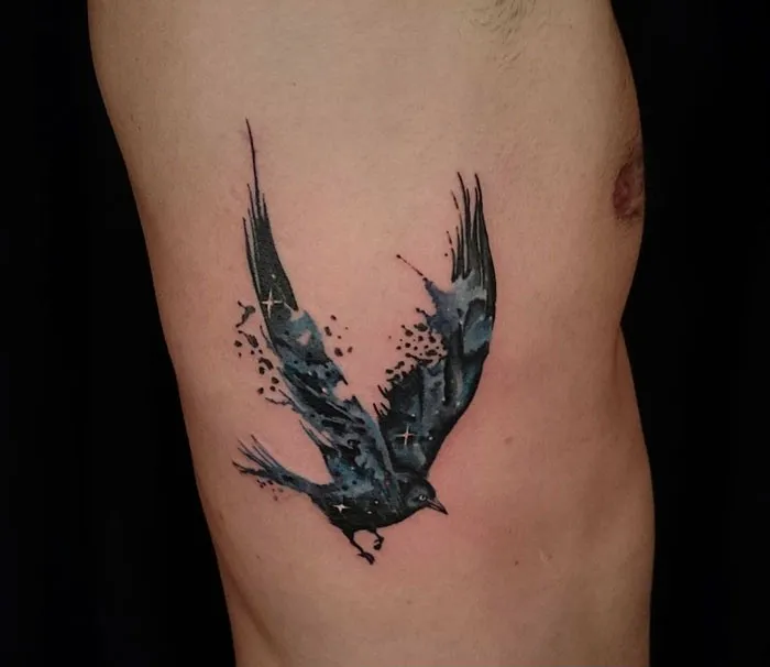Watercolor style small bird tattoo on side, featuring a flying blue-black bird with dynamic brush strokes.