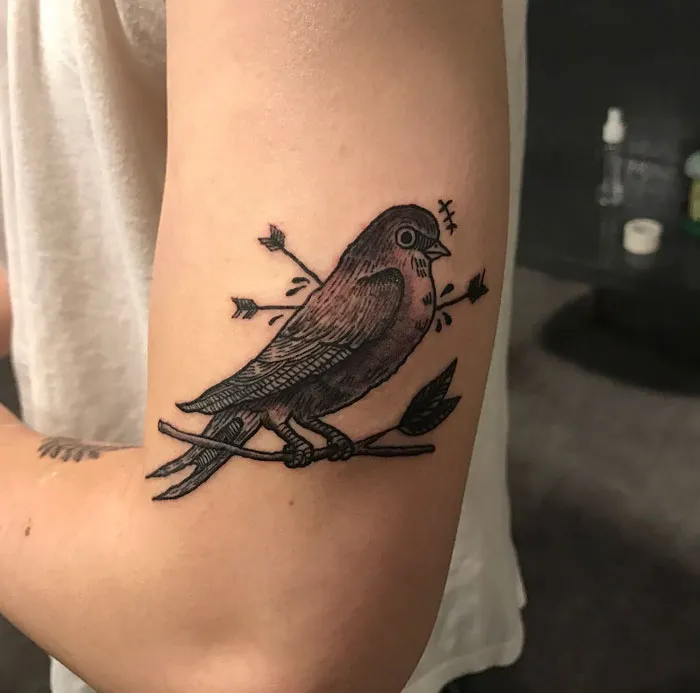Unique small bird tattoo with arrows, inked on upper arm.