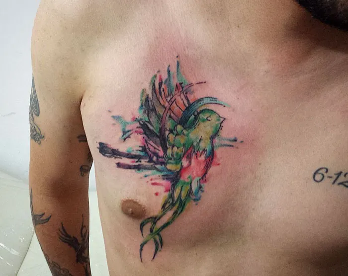 Colorful small bird tattoo on a man's chest, featuring vibrant watercolor style.