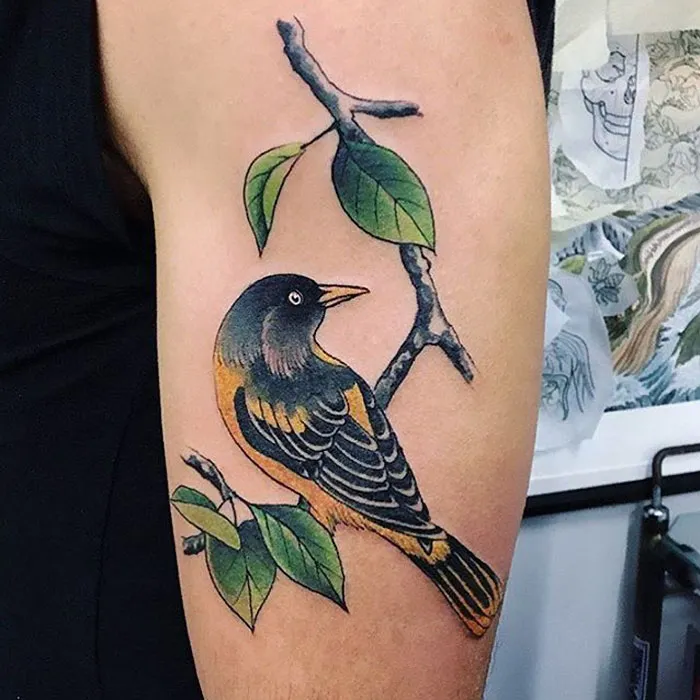 Colorful small bird tattoo on an arm, featuring intricate details with a branch and leaves.