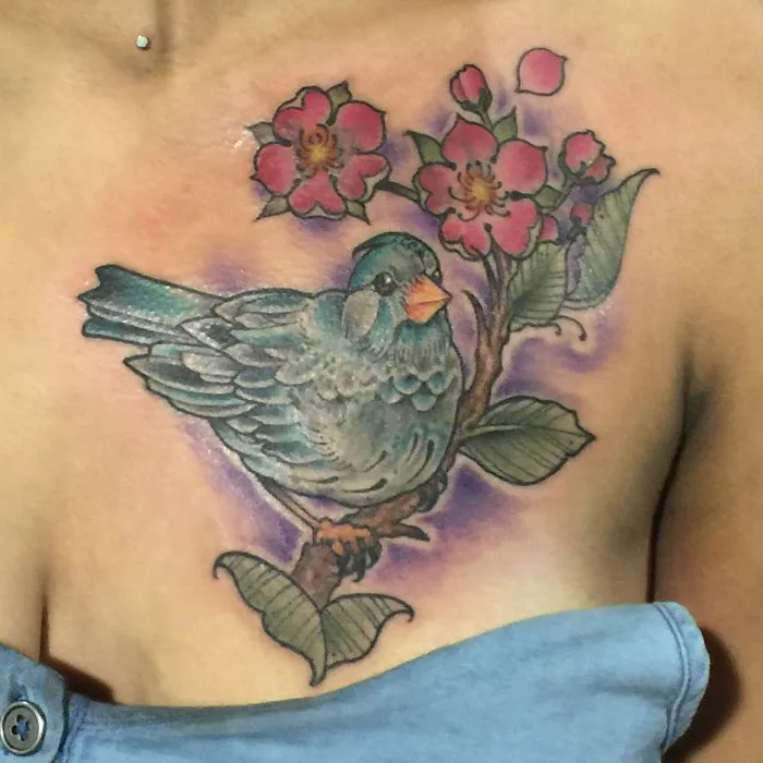Colorful small bird tattoo design featuring flowers on a person's shoulder.