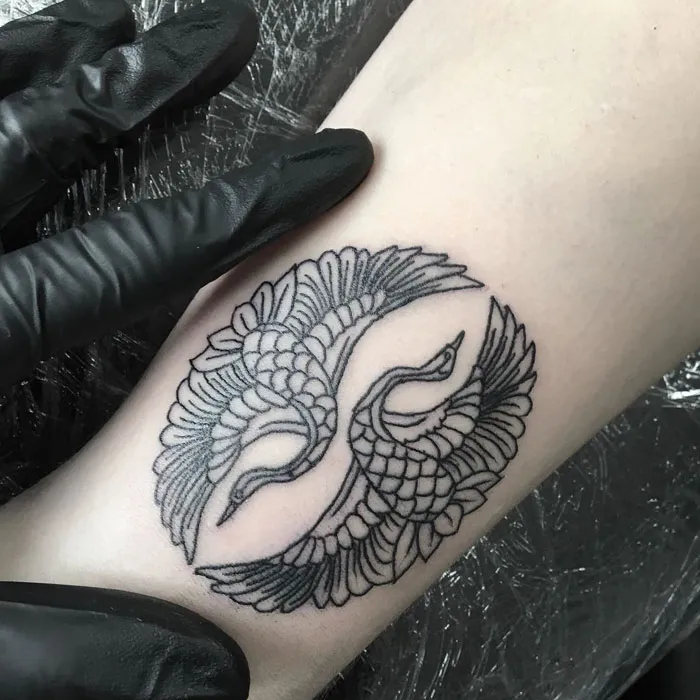 Intricate small bird tattoo design with two entwined cranes on forearm, displayed by gloved hand.