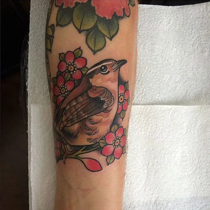 Colorful small bird tattoo among flowers on forearm, showcasing unique tattoo artistry.