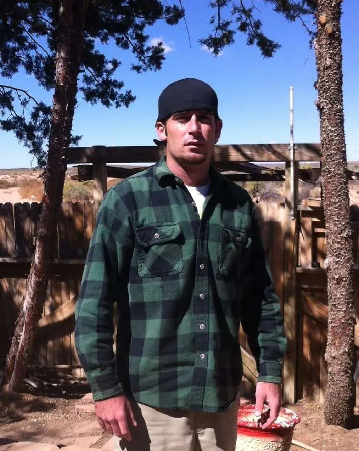 Man standing outdoors between trees wearing a green plaid shirt and backward black cap showing effects of stopping drinking.