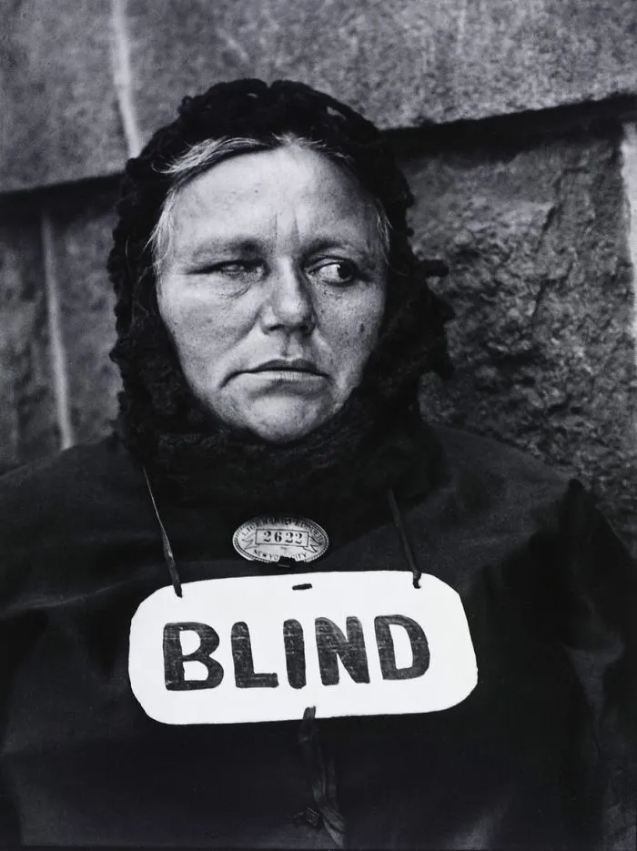 Woman in a headscarf with "BLIND" sign, from a famous historical photo, against a textured wall.