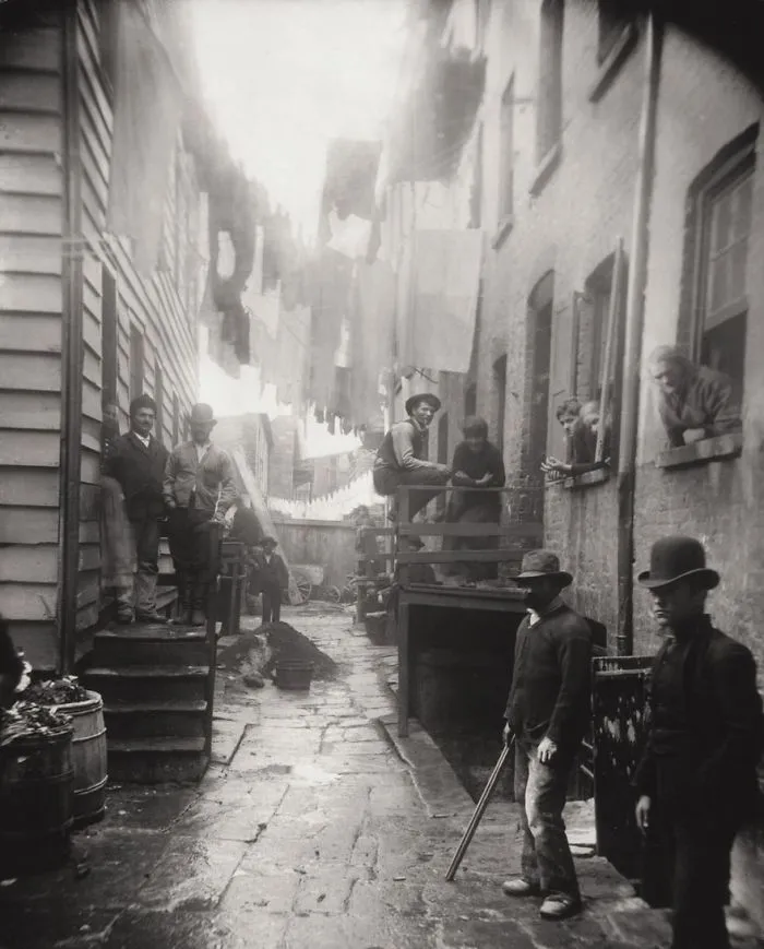 Historic photo of people in a narrow alley, wearing hats and traditional clothing, with laundry hanging above.