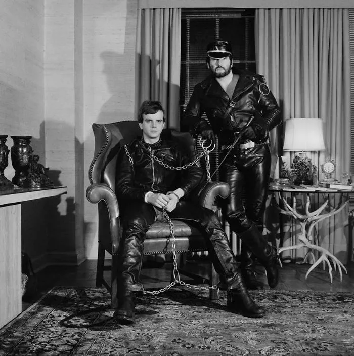 Two men in leather outfits, one seated and one standing, in a room with vintage decor, representing famous photos in history.