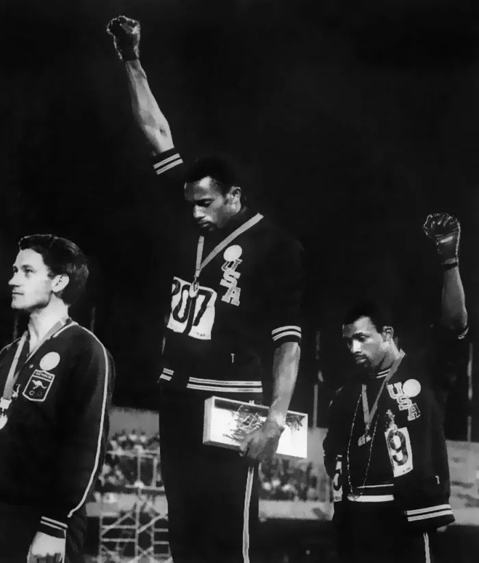 Athletes on a podium, two raising fists in a famous protest during an Olympic medal ceremony.