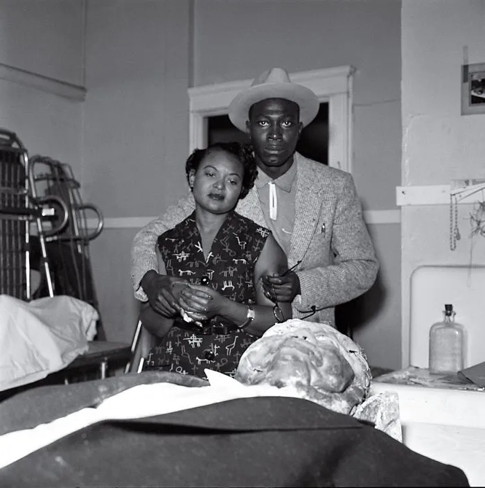 A historical photo of a couple in a somber setting, depicting a poignant moment with an embalmed body in the foreground.