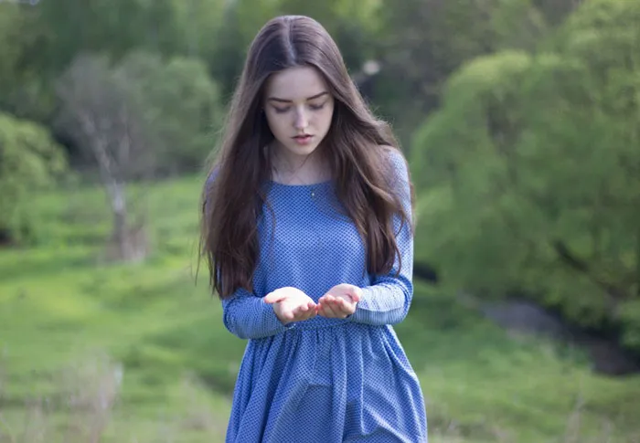 Young woman in a blue dress stands in a lush green field, looking down at her hands, showcasing beauty before Photoshop.