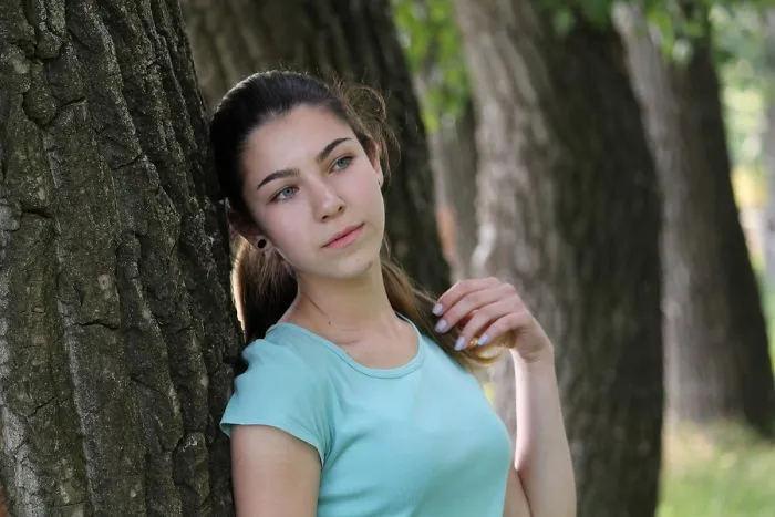 Woman in a park, leaning against a tree, wearing a light blue shirt, representing truth behind beautiful photos.