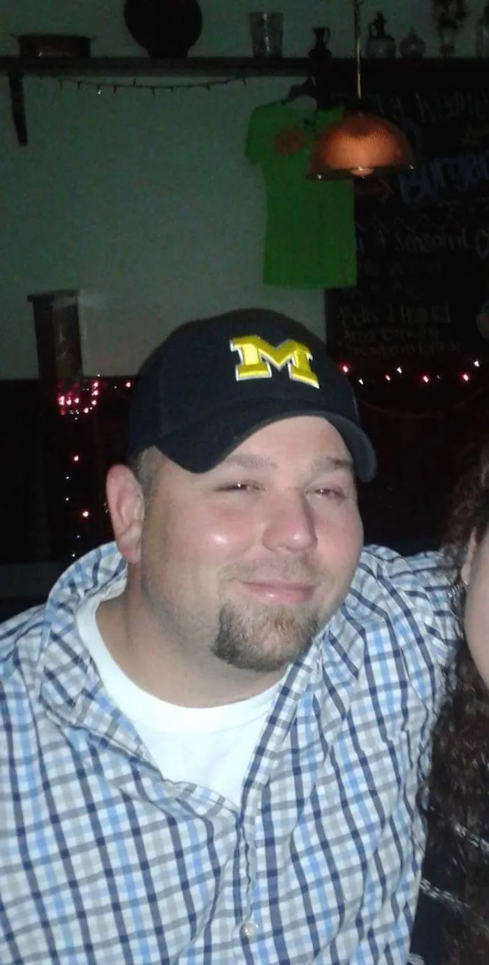 Man wearing a Michigan cap and checkered shirt smiling in a dimly lit bar, illustrating effects of stopping drinking.