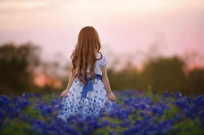A girl in a blue floral dress stands in a field of flowers at sunset, showcasing beauty before Photoshop.