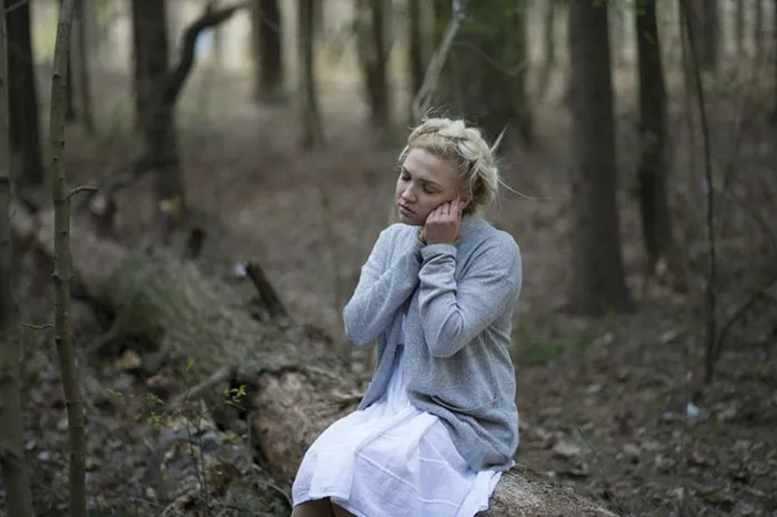 Woman in the woods, sitting on a log, before Photoshop transformation, wearing a gray sweater and a white dress.