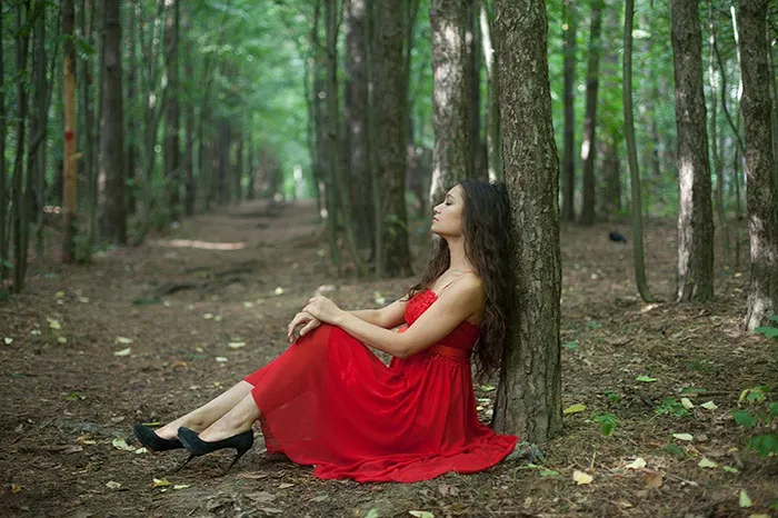 Woman in a red dress sitting against a tree in a wooded area, highlighting Photoshop's impact on beautiful photos.