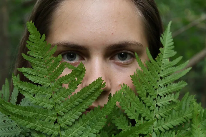 Woman's face partially obscured by green ferns, highlighting natural beauty before photoshop.