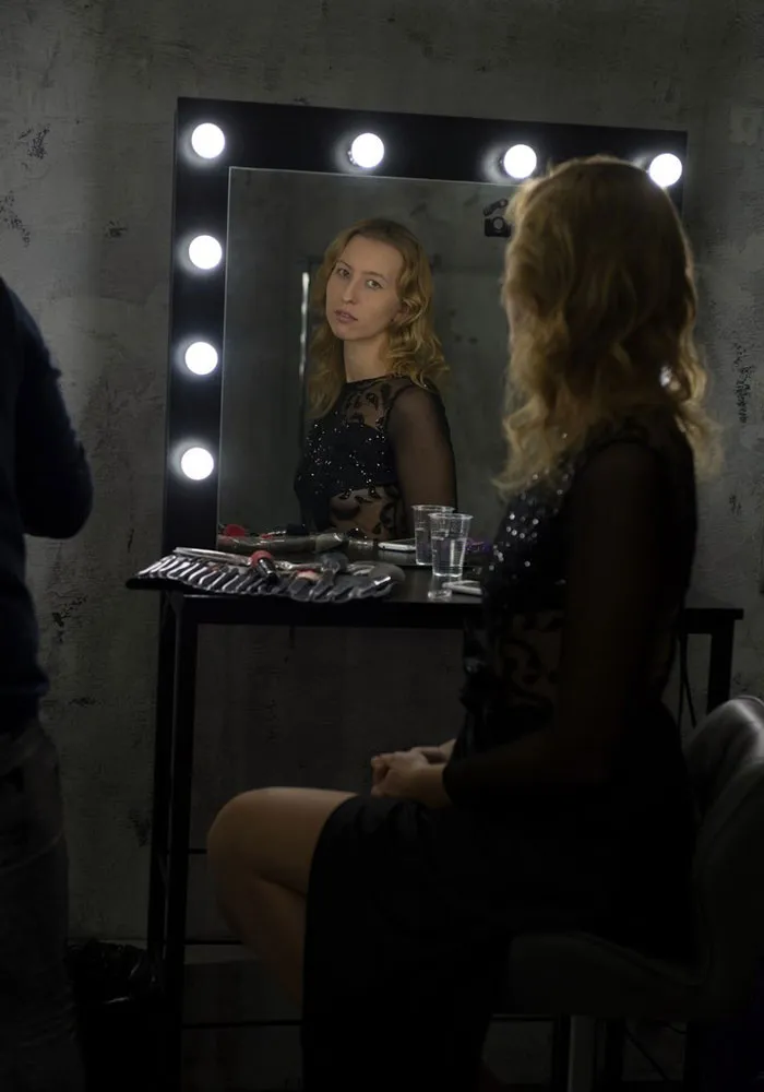Woman in front of a lit mirror, before and after Photoshop, dressed in a black dress with makeup tools on the table.