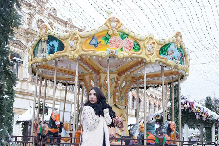 Woman in winter clothing standing in front of a colorful carousel, revealing Photoshop's impact on beautiful photo aesthetics.