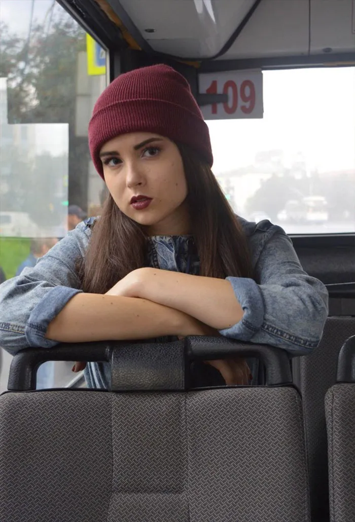 Woman in a denim jacket and maroon beanie sitting on a bus seat, natural beauty before Photoshop adjustments.