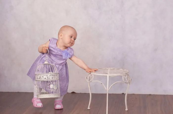 Baby in a purple dress holding a birdcage, reaching for a small table, illustrating before and after Photoshop transformations.
