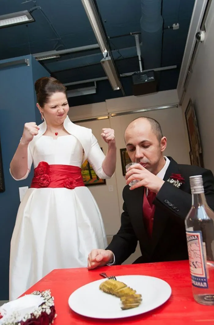 Bride in white dress stands playfully triumphing as groom in tux drinks, highlighting Photoshop contrast in beautiful photos.