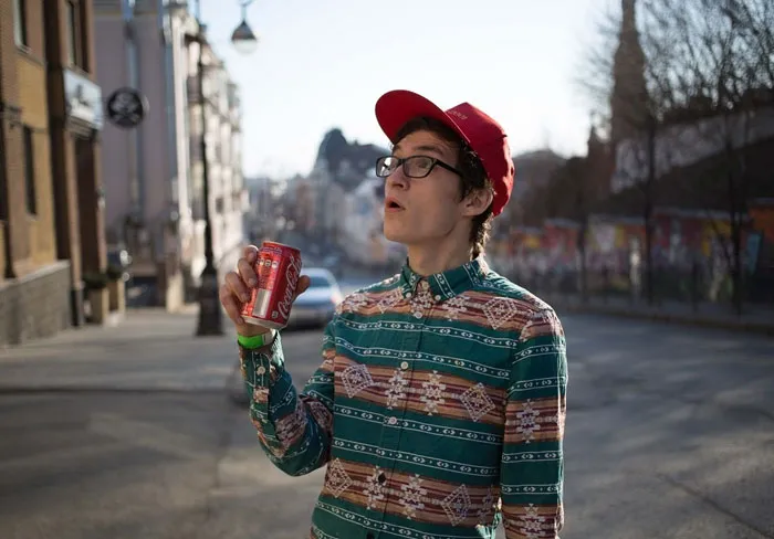 Person in a patterned shirt and red cap, holding a soda can, standing on a sunny street before Photoshop.