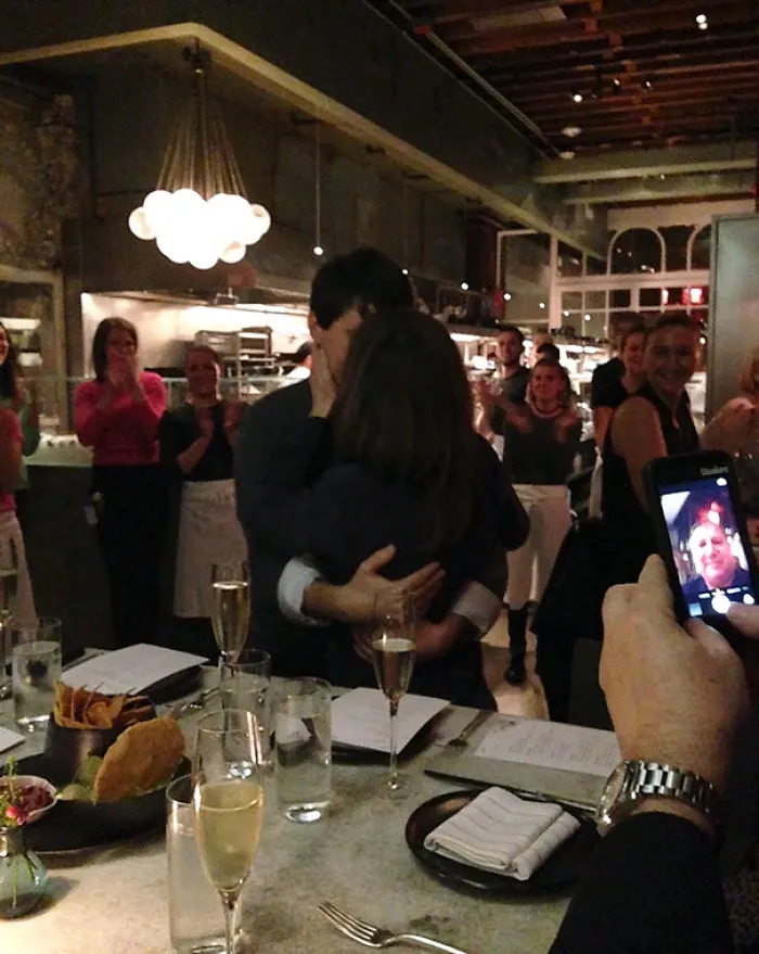 A couple embraces in a lively restaurant as a person captures a photo, creating a "when you see it" moment.