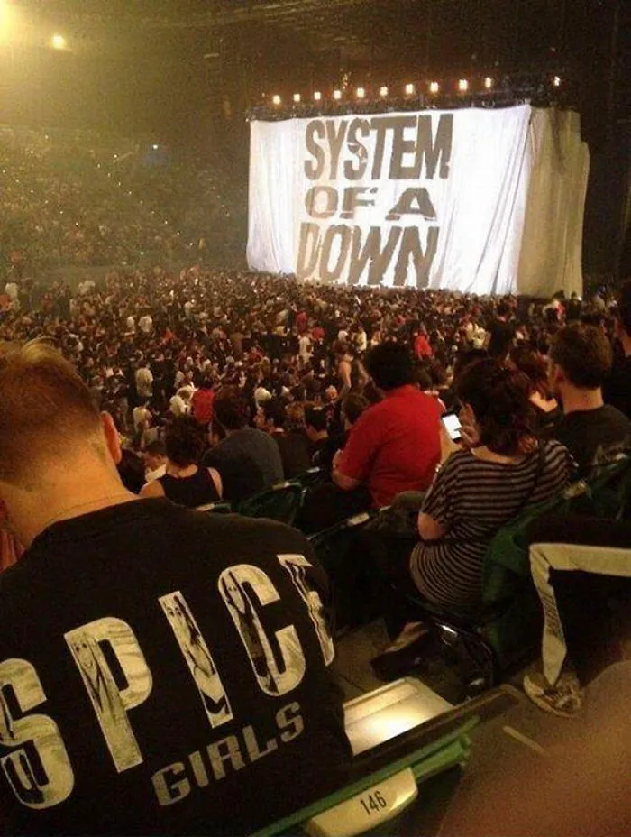 Concert crowd with "System of a Down" curtain, person in foreground wearing a "Spice Girls" shirt, unexpected contrast.