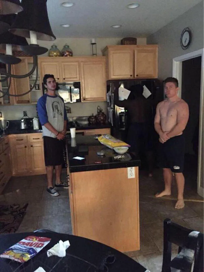 Two men in a kitchen with a mysterious figure by the fridge, a classic "when you see it" moment.
