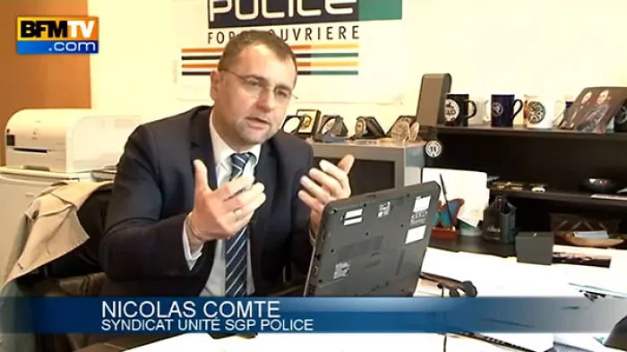 Man in office setting discussing with expressive hand gestures, police banner in background.
