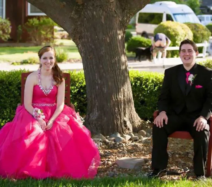 Couple in formal attire sitting near a tree, with a humorous background detail that surprises upon closer inspection.