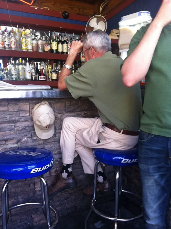 Man at bar with odd positioning, wearing green shirt and beige pants, sitting on a blue Budweiser stool.