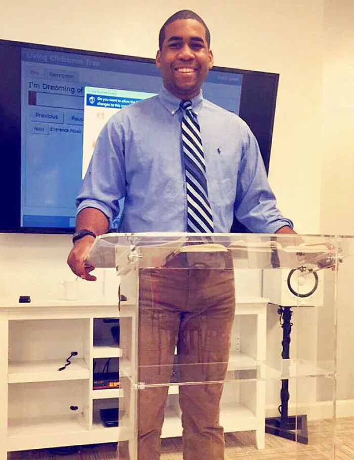 Man smiling behind a clear podium, wearing a blue shirt and striped tie, with a presentation screen in the background.