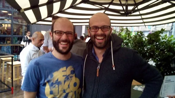Two smiling men with glasses in a cafe, appearing as real-life doppelgangers.