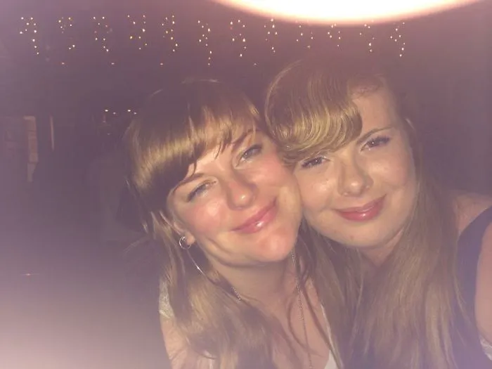 Two women smiling closely, looking like real-life doppelgangers, illuminated by party lights.