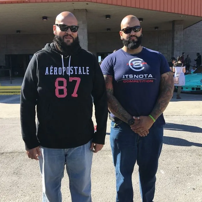 Two men with similar build and beards, wearing sunglasses, appear as real-life doppelgangers outside a building.