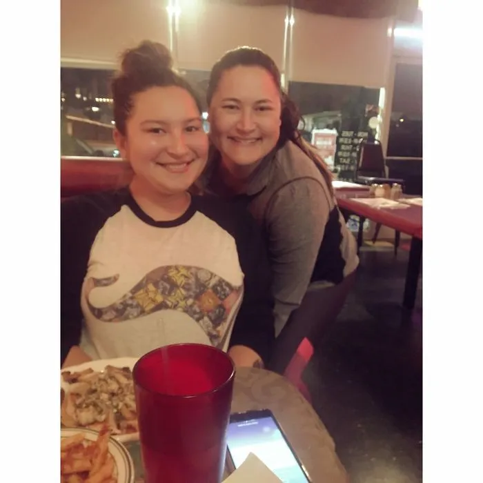 Two women smiling, each resembling real-life doppelgangers, sitting at a restaurant table with food and drinks.