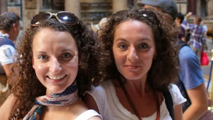 Two women with curly hair and sunglasses meet, resembling real-life doppelgangers, smiling in an outdoor setting.