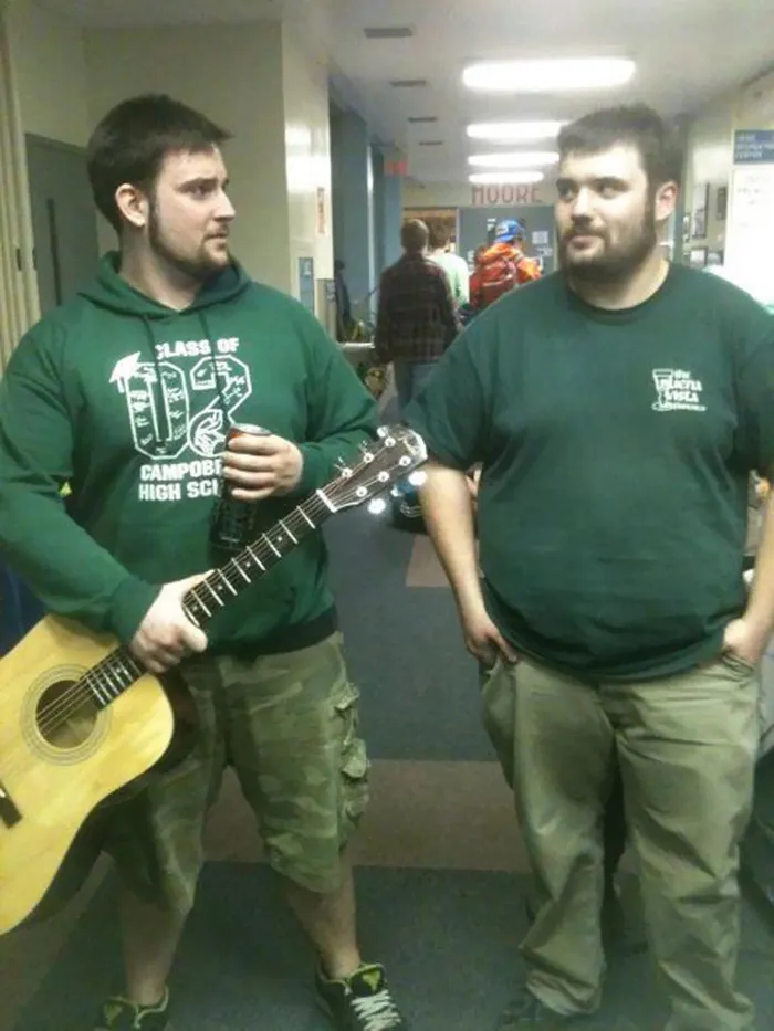 Two men in green shirts and shorts, standing in a hallway, looking alike like real-life doppelgangers.