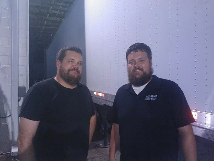 Two men meeting their real-life doppelgangers, standing next to a truck in casual attire.